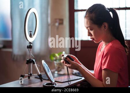 Vue latérale d'une jeune femme asiatique concentrée assise à table avec des cosmétiques et des outils pour le tournage vidéo et la vérification vidéo sur l'appareil photo tout en créant du contenu pour le blog de beauté à la maison Banque D'Images
