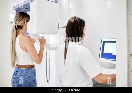 Vue arrière d'une femme dentiste utilisant un ordinateur tout en effectuant un scan à rayons X à une femme blonde dans une clinique dentaire contemporaine Banque D'Images