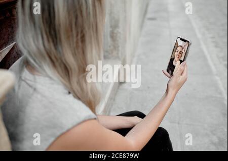 Coupe courte grand angle pour une jeune femme méconnue dans une ambiance décontractée vêtements assis sur des marches de pierre dans la rue et en regardant la vidéo conversation avec un ami via un smartphone Banque D'Images