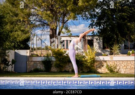 Vue latérale du soutien-gorge de sport et des leggings fins pour femme se tenir sur un tapis près de la piscine et pratiquer le yoga à Urdhva Hastasana Banque D'Images