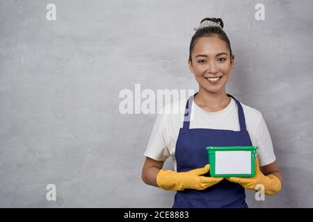 Portrait d'une jeune femme souriante, femme de ménage en uniforme et gants en caoutchouc jaune tenant une boîte en plastique vert avec des capsules de lavage tout en se tenant contre un mur gris. Le concept de ménage et de ménage Banque D'Images