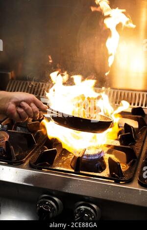 Un chef cuisinier écourté et méconnu lanche des crevettes dans du flambe au-dessus du gaz faire cuire au feu de bois tout en cuisinant au restaurant Banque D'Images