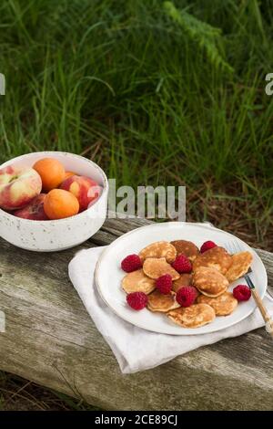 Assiette avec crêpes fraîchement cuites placée sur un banc en bois près bols avec fruits pour le petit déjeuner à la campagne Banque D'Images