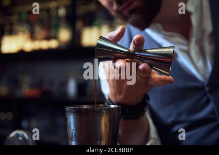 Jeune barman professionnel non reconnaissable, court, versant un cocktail d'alcool du puzzle dans un shaker métallique sur le comptoir pour faire des boissons bar Banque D'Images