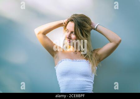 Jeune femme pleine de confiance en tenue d'été se touchant la tête pendant debout contre le mur blanc le jour ensoleillé et regardant loin Banque D'Images