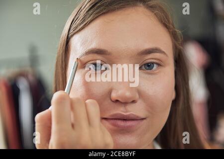 Un maquilleur axé sur la récolte qui fait des sourcils sur une jeune belle femme client regardant la caméra pendant son travail dans un studio contemporain Banque D'Images