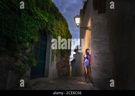 Calme jeune femme ethnique dans des vêtements colorés et décontractés mur en pierre d'un bâtiment ancien tout en explorant l'ancienne rue de Tossa de Mar commune en Espagne en soirée d'été Banque D'Images