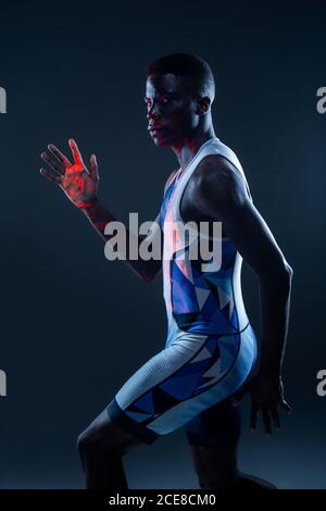 Vue latérale d'un sportif afro-américain sérieux et sportif en couleurs active wear pour des mouvements rapides en studio sombre avec éclairage au néon rouge Banque D'Images
