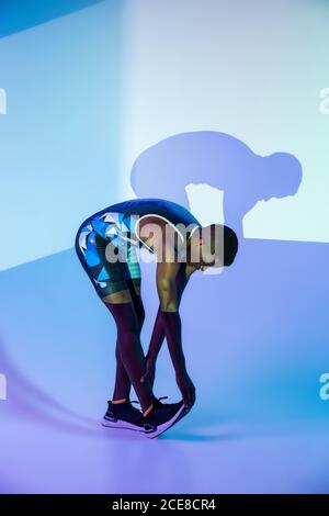 Vue latérale complète du corps d'un homme afro-américain déterminé sprinter en costume de sport se pliant vers l'avant et étirant les jambes avant commencez en studio avec un éclairage au néon bleu Banque D'Images