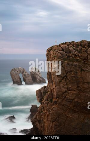 Vue latérale silhouette distante de la personne se tenant sur le bord de falaise rocheuse près de mouettes et de rochers en pleine ascension mer contre un magnifique ciel de couleur lilas à Santander Espagne Banque D'Images