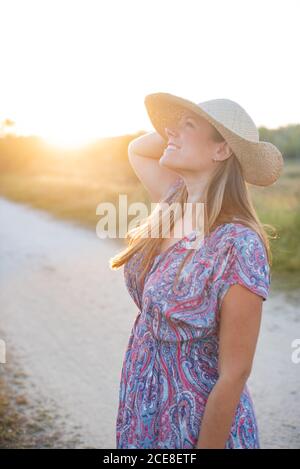 Charmante femme en robe élégante et chapeau de paille debout route de sable dans la zone rurale et profiter du coucher du soleil pendant le week-end Banque D'Images