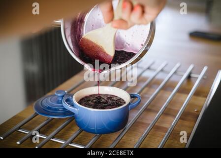 Crop anonyme femme au foyer avec une cuillère en bois verser savoureux fraîchement préparé confiture de myrtilles de la poêle dans la casserole de céramique pendant la préparation du petit déjeuner à la maison Banque D'Images