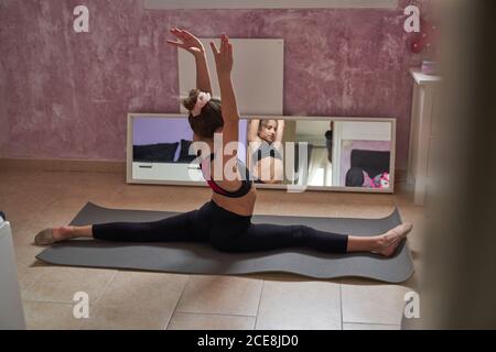 Vue arrière d'une jeune fille mince assise sur un tapis faisant se fend avec les bras levés pratiquant la gymnastique devant le miroir Banque D'Images