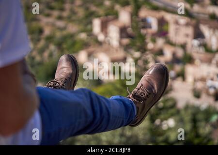 Vue latérale du petit randonneur anonyme en Jean tendance et des bottes assises sur le bord de la montagne contre la vieille pierre ville et profiter des vacances d'été Banque D'Images