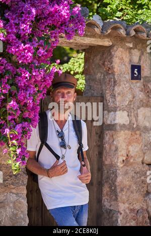 Taille basse sérieuse d'âge moyen homme dans des vêtements tendance avec sac à dos debout près d'un mur de pierre aux intempéries avec porte en bois et fleurs plantes lors de la visite de la vieille ville Banque D'Images