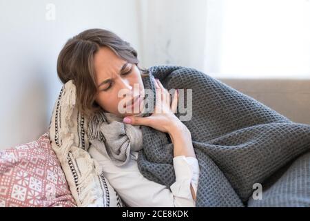 Une jeune femme malade couché et tremblante, enveloppée de couvre-maillot et de foulard, tente de se réchauffer, ayant des symptômes de grippe Banque D'Images