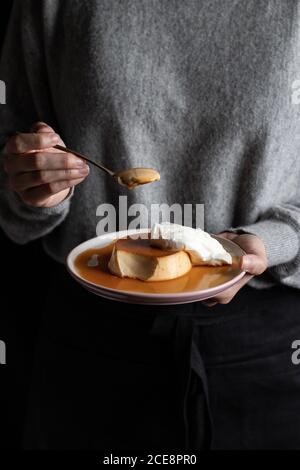 une femelle méconnaissable mange une délicieuse crème au caramel placée sur l'assiette avec crème fouettée sur fond noir Banque D'Images