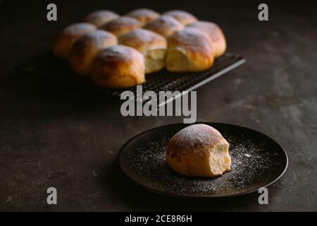 De délicieux petits pains ronds frais à texture douce et surface dorée décorée de sucre en poudre sur la grille de refroidissement et tableau sur fond noir flou Banque D'Images