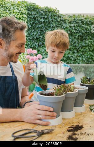 Homme gai en tablier et garçon en utilisant la truelle de jardinage pendant arrosage du sol des plantes en pots cactus plantules debout près de la table derrière des buissons verts luxuriants Banque D'Images