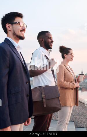 Vue latérale des étudiants en affaires multiethniques souriants qui se tiennent debout et contempler le paysage urbain tout en planifiant l'avenir Banque D'Images