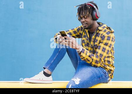 jeune africain avec casque assis sur un mur jaune en consultant son smartphone avec un mur bleu en arrière-plan, technologie et concept de style de vie, espace de copie pour le texte Banque D'Images