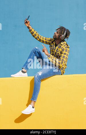 jeune homme noir assis sur un mur jaune avec un casque pour passer un appel vidéo avec son smartphone avec un mur bleu en arrière-plan, concept de technologie et de style de vie, espace de copie pour le texte Banque D'Images