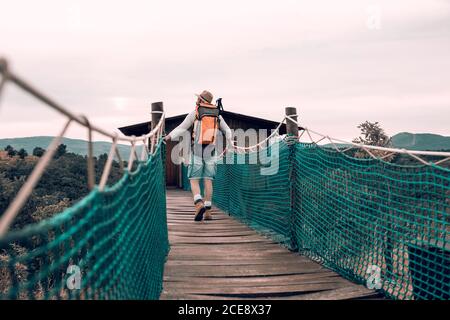 Vue arrière de contenu inconnu mâle touriste avec marche à dos le long du pont suspendu et en admirant le paysage étonnant pendant les vacances à été Banque D'Images