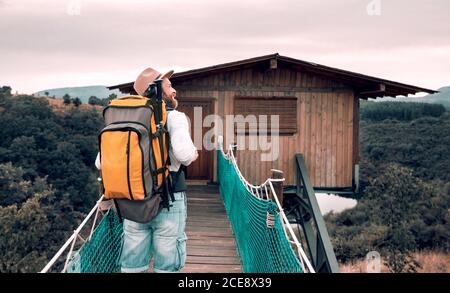 Vue arrière de contenu inconnu mâle touriste avec marche à dos le long du pont suspendu et en admirant le paysage étonnant pendant les vacances à été Banque D'Images