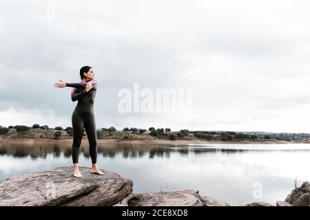 Femme pleine hauteur pieds nus en combinaison noire pour un échauffement les articulations des épaules tout en étirant les bras de côté sur une rive paisible de lac en couvert Banque D'Images