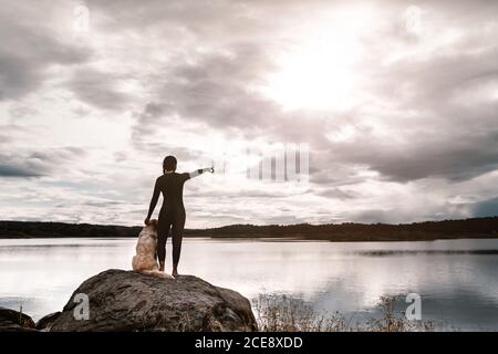 Vue arrière de la femme anonyme en combinaison noire debout avec le chien sur la pierre et pointant vers l'extérieur tout en observant la nature du lac Banque D'Images