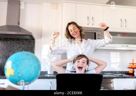 Femme gaie dans une tenue habillée debout avec les bras relevés derrière un garçon positif dans un casque touchant la tête tout en regardant le netbook et étudiant à la maison près du globe Banque D'Images
