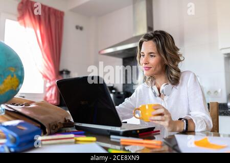 Une femme adulte gaie dans un casque assis à la table avec une tasse de boisson chaude devant le netbook pendant le chat vidéo près de la papeterie colorée dans la cuisine légère Banque D'Images