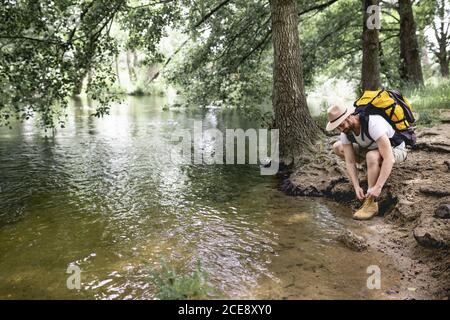 jeune homme randonnée avec sac à dos jaune et chapeau lui attacher lacets sur la rive d'un lac rempli d'arbres Banque D'Images