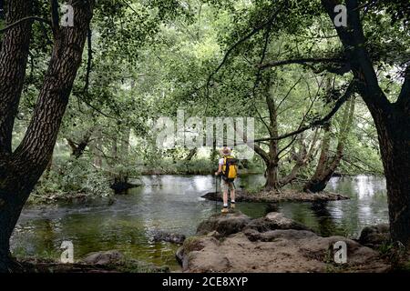 jeune homme faisant un sentier de randonnée avec son sac à dos jaune et chapeau sur sa tête par un lac avec beaucoup arbres et zones naturelles qui donnent sur le paysage Banque D'Images