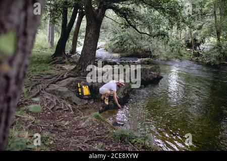 jeune randonneur avec sac à dos jaune et chapeau de repos et prendre de l'eau sur la rive d'un lac plein de arbres verdoyants Banque D'Images