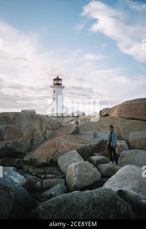 Vue latérale de l'homme anonyme debout sur le chemin de poney et En regardant le phare de Peggys Cove contre le ciel nuageux et ensoleillé Canada Banque D'Images