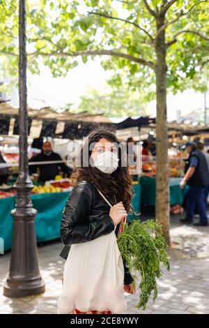 Vue latérale de la femme calme portant un masque de protection du visage avec sac à provisions écologique choisir des légumes frais sur un marqueur local Banque D'Images