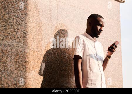 Jeune homme noir concentré en chemise blanche debout au mur à l'extérieur et en lisant des sms sur le téléphone ou en vérifiant le courrier Banque D'Images