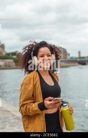 Femme athlète en tenue active et casque tenant un téléphone portable après entraînement en se tenant sur une bankment près de la rivière de la ville Banque D'Images