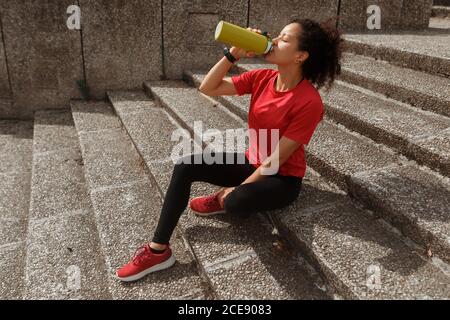 Vue latérale d'une athlète féminine de sport et une montre intelligente pour boire de l'eau à partir de thermos tout en restant avec les yeux fermés sur l'escalier après l'entraînement à la lumière du soleil Banque D'Images