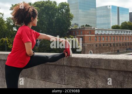 Vue latérale d'une sportive ethnique anonyme en tenue active avec une coupe d'Afro sur un pont en pierre pendant l'entraînement devant les bâtiments de la ville en plein jour Banque D'Images