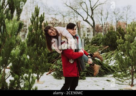 Un jeune couple se berce, s'amusant en hiver sur le fond du marché des arbres de Noël dans la ville. Une jeune femme sur le dos de son petit ami rit et sourit. Banque D'Images