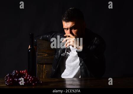 Un homme à la barbe boit un verre de vin sur fond marron foncé. Le sommelier goûte des boissons chères. Concept de dégustation de vins et de dégustation. Le Degustator au visage plein de confiance se trouve à côté d'une bouteille de vin et de raisins foncés Banque D'Images