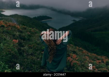 Vue arrière de la jeune femme se tenant sur la colline avec l'herbe verte et regardant le beau lac en dessous Banque D'Images