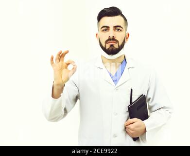 Le médecin avec la barbe tient un carnet pour les antécédents de maladie et affiche le signe ok. Homme au visage sérieux sous un manteau médical blanc. Concept de traitement réussi. Guy dans le masque chirurgical isolé sur fond blanc Banque D'Images