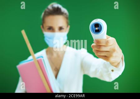 La vie durant la pandémie du coronavirus. Gros plan sur la femme enseignante en blouse blanche avec masque médical mesure la température avec thermomètre numérique sur le chalkboa Banque D'Images