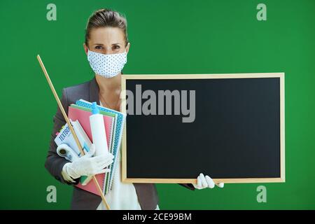 femme moderne enseignante d'école en blouse blanche avec masque médical, gants en caoutchouc, thermomètre numérique, manuel et assainisseur montrant blanc tableau iso Banque D'Images