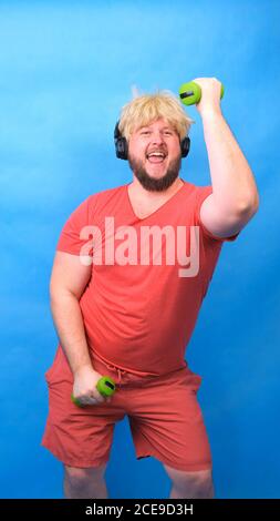 Drôle de chubby freak homme dans le casque et une perruque dans un t-shirt rose est engagé dans les haltères vertes et rires sur un fond bleu, vertical Banque D'Images