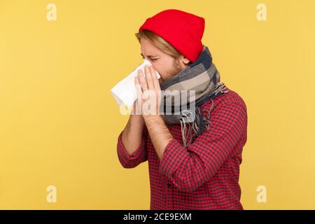 Homme malade en chapeau et foulard chaud toussant le tissu éternuant, se sentant mal avec l'écoulement nasal, mal de gorge, souffrant d'allergie ou de symptômes de grippe saisonnière Banque D'Images