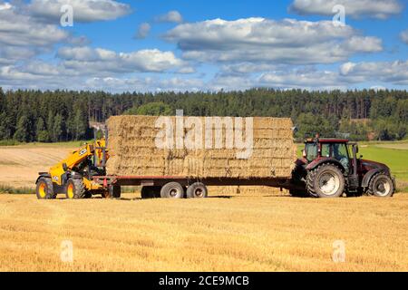 Le téléchargeur JCB T4 empilant des balles de paille sur un tracteur Valtra a tiré une remorque agricole sur un terrain de chaume par beau temps. Salo, Finlande. 16 août 2020. Banque D'Images